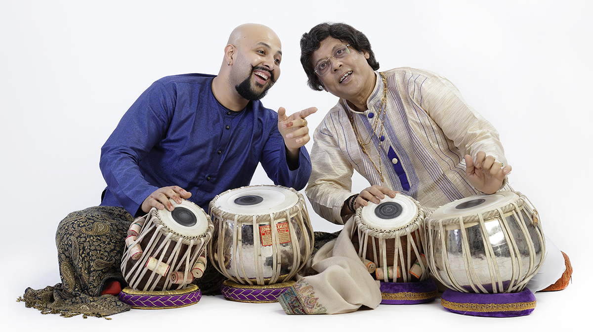 A bald Indian man in a blue shirt leans towards an older man. Both rest their hands on tablas, a type of South Asian drum